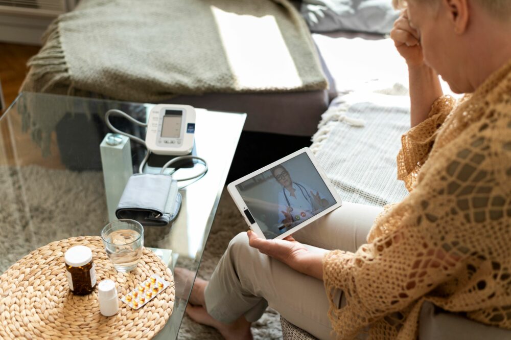 Femme senior assise à domicile en consultation de télémédecine sur tablette, sur la table basse devant elle sont posés : un tensiomètre et des médicaments